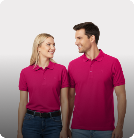 Man and a woman stand side-by-side, both wearing matching bright pink polo shirts and blue jeans, looking at each other and smiling