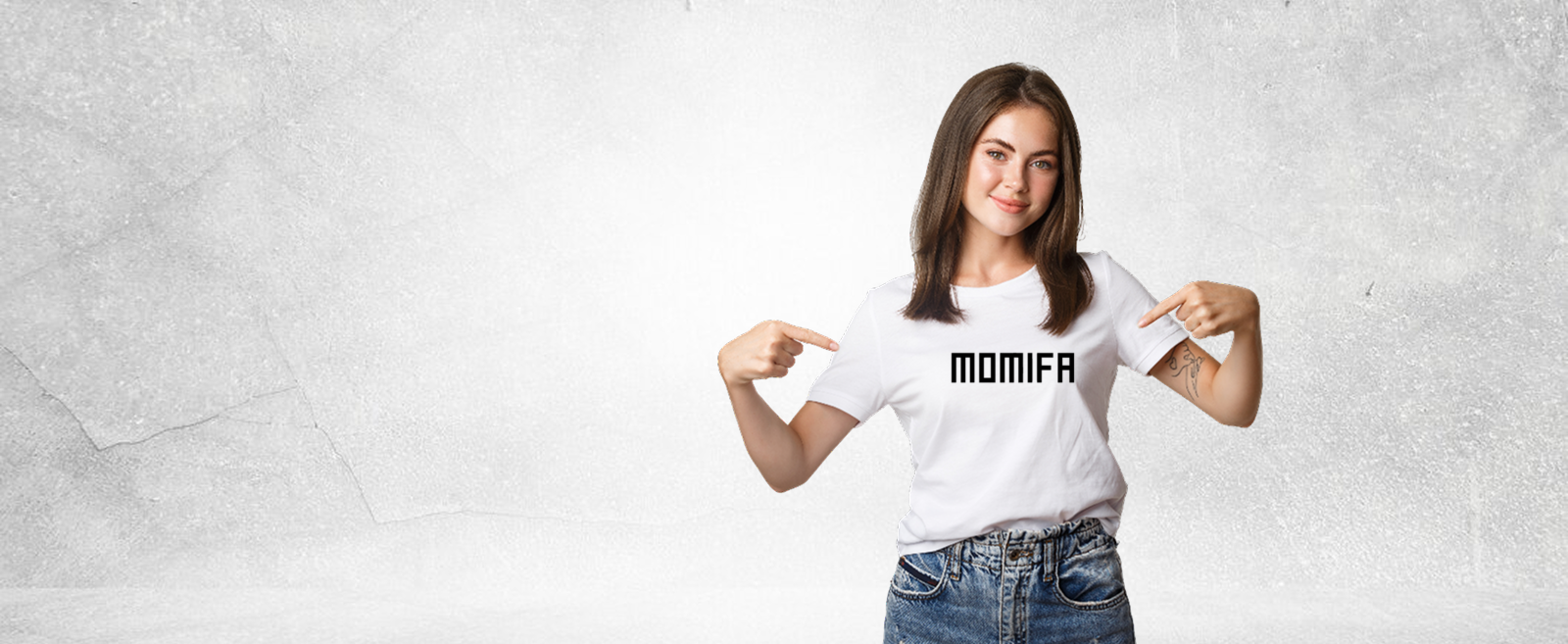 Smiling woman with long brown hair wearing a white t-shirt with the word "MOMIFA" printed on the chest, pointing to the text with both hands.