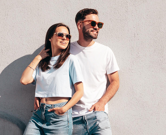 smiling man and woman wearing sunglasses, t-shirts, and jeans, posing together in front of a textured gray wall.