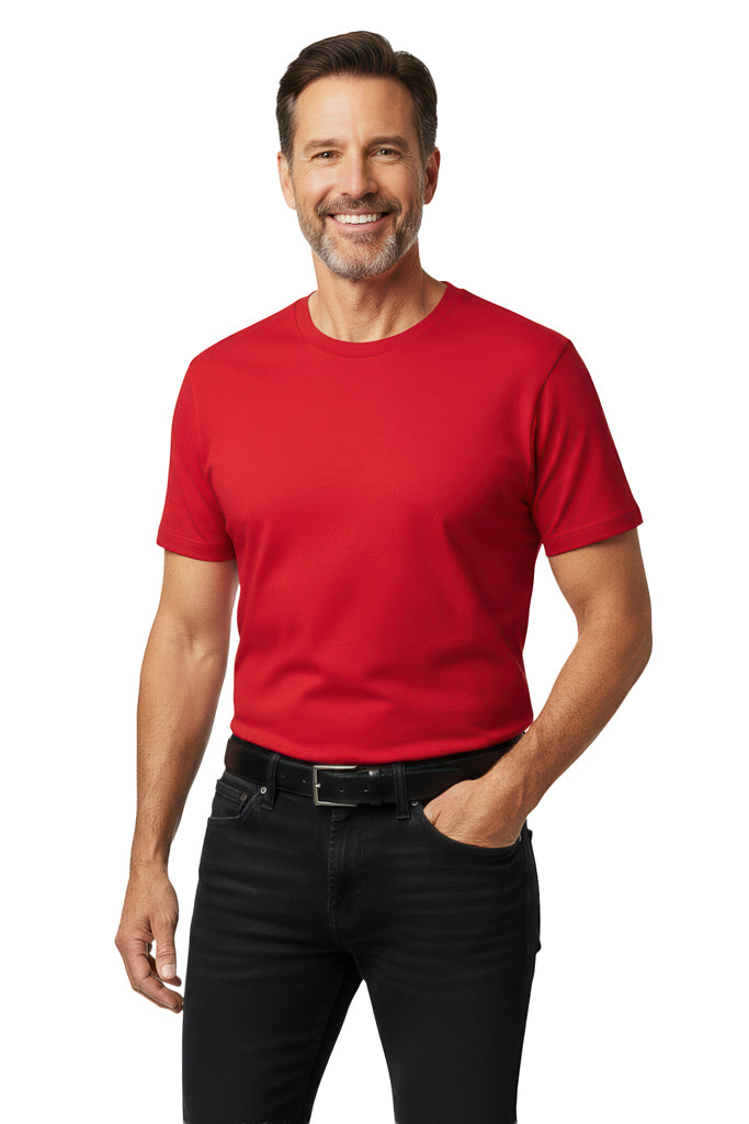 Man with a beard and dark hair, wearing a red t-shirt and black jeans, smiles while standing with a hand in his pocket.